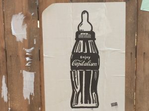 This image shows a poster of a Coke bottle with the phrase "Enjoy Capitalism" on it. The bottle also has a pacifier on top.